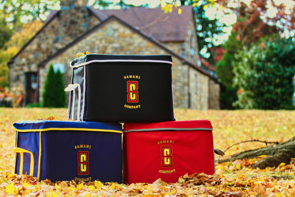 Three Samani Company storage bags are stacked in a pyramid formation outdoors on fallen autumn leaves. The top bag is black with white trim, while the bags below are red with gray trim on the bottom left and navy blue with yellow trim on the bottom right. All bags feature the Samani Company logo.